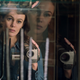 A woman holds a video camera while staring into a glass case looking at tuning forks, with reflections of her image on either side.